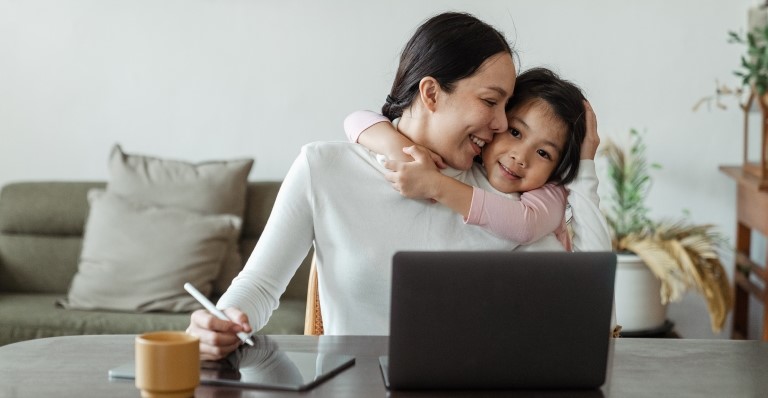 A child embracing her mother as she is working on her computer