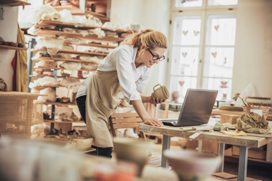 Une femme dans son atelier tient une pièce de poterie tout en regardant son ordinateur