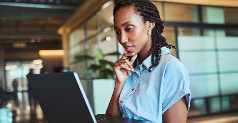 Thoughtful young Black professional with braided hair studies laptop, chin resting on hand, in open-plan office