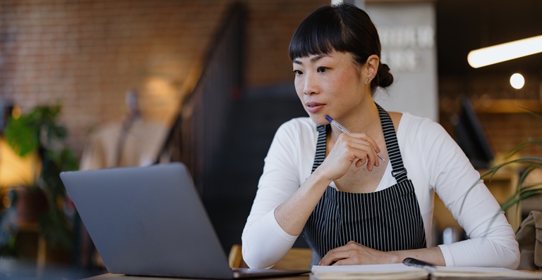 Upscale coffee shop owner wearing apron sits thoughtfully with laptop and notebook, planning menu.