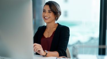 A businesswoman smiling while working on her computer at a desk in an office setting.