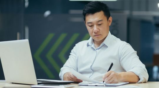 Un homme d’affaires, assis à un bureau, signe des documents, un ordinateur portable se trouvant posé devant lui.