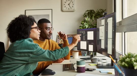 Un homme et une femme, assis à un bureau, regardent les graphiques s’affichant sur deux moniteurs; la femme pointe les écrans avec un stylo.