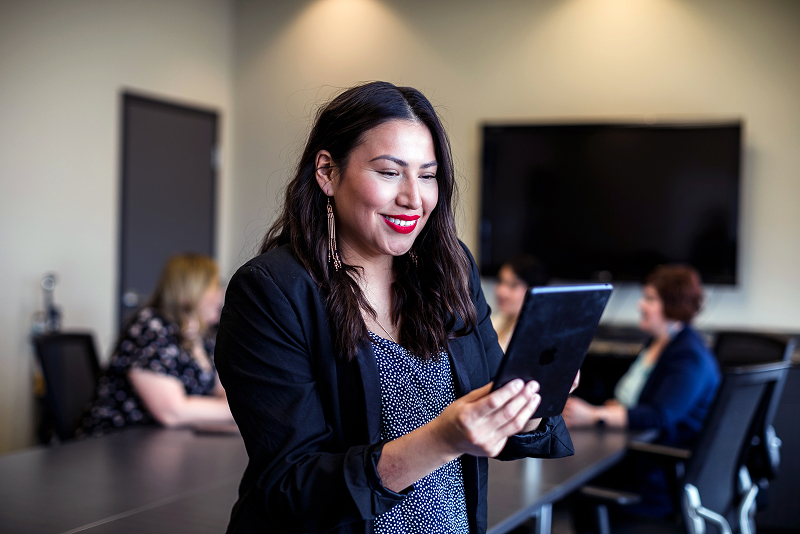  Person holding a tablet in a modern conference room with colleagues seated at a table and a large screen on the wall.