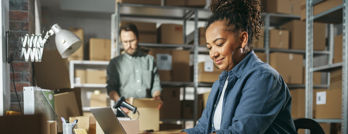 Deux employés d'entrepôt : l'un utilise un ordinateur portable tandis que l'autre vérifie des cartons dans une pièce remplie de cartons rangés sur des étagères.
