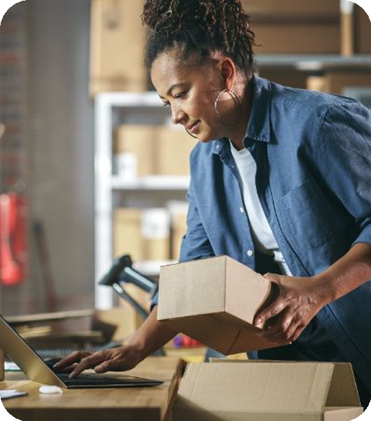 Woman in a warehouse using a laptop and handling a box.