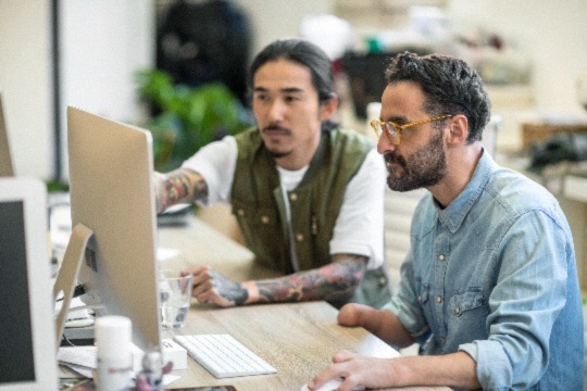 A man is pointing toward a computer monitor to indicate something to another man