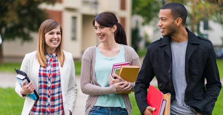 Three young adults walking on a campus with books in their arms.