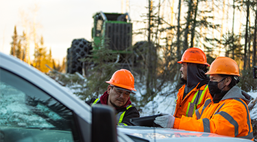 Trois hommes portant casque et veste, et se tenant près d’un véhicule, regardent une tablette. Derrière eux, un tracteur vert.