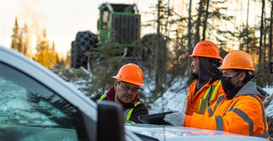 Trois hommes portant casque et veste, et se tenant près d’un véhicule, regardent une tablette. Derrière eux, un tracteur vert.