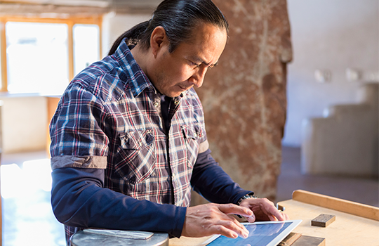 A man in a checkered shirt is working at a desk on a tablet.