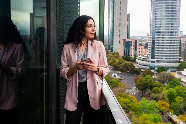 Personne debout sur un balcon tenant un téléphone intelligent, avec en arrière-plan un paysage urbain composé d’immeubles modernes et d’arbres verts.