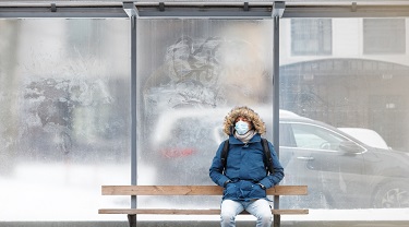 Homme malade avec un capuchon assis seul sur un banc de transport public, portant un masque facial de protection