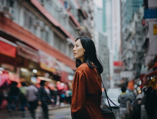 A woman stands on a busy street, surrounded by tall buildings and looking pensive.