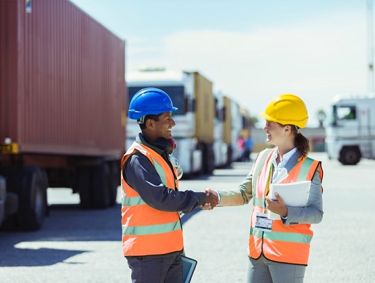 A man and a woman wearing construction hats shake hands and smile.