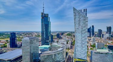 Un paysage urbain animé de Varsovie, en Pologne, avec plusieurs gratte-ciels modernes sous un ciel bleu clair. Le plus haut bâtiment au centre possède une façade en verre élégante, tandis qu’un autre bâtiment important à droite présente un design unique et angulaire avec des bandes horizontales.