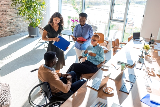 Group of professionals of varying ages, ethnicities, and abilities engaging in conversation in a modern office