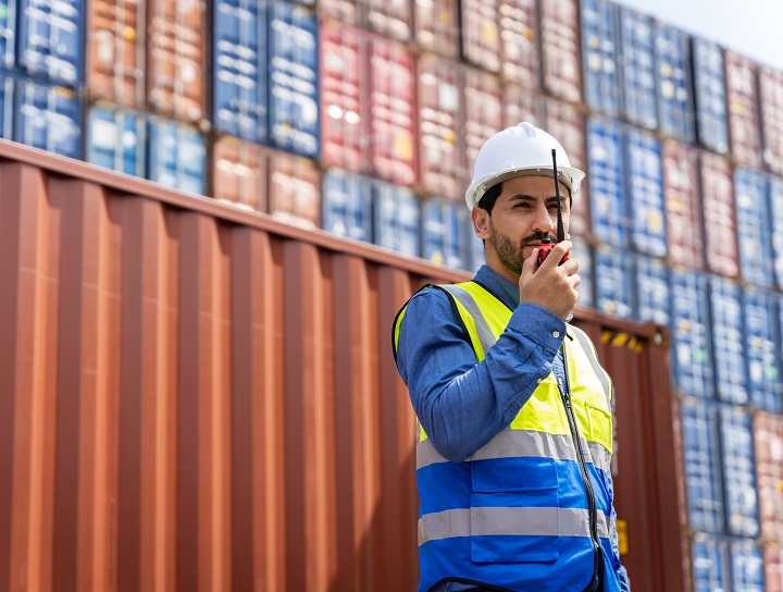 Male worker talks on handheld radio with shipping containers in the background.