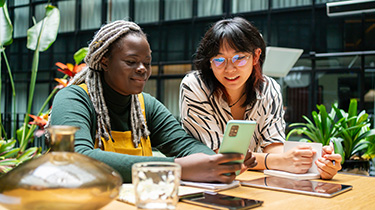Image de deux femmes avec téléphone cellulaire, cahier de notes et tablettes, épanouies dans un milieu de travail inclusif et équitable.