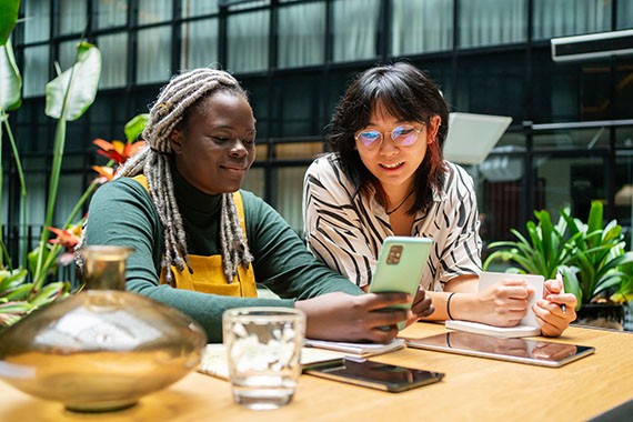 Image of two women with mobile phone, notepad and tablets flourishing in an inclusive, equitable workspace.