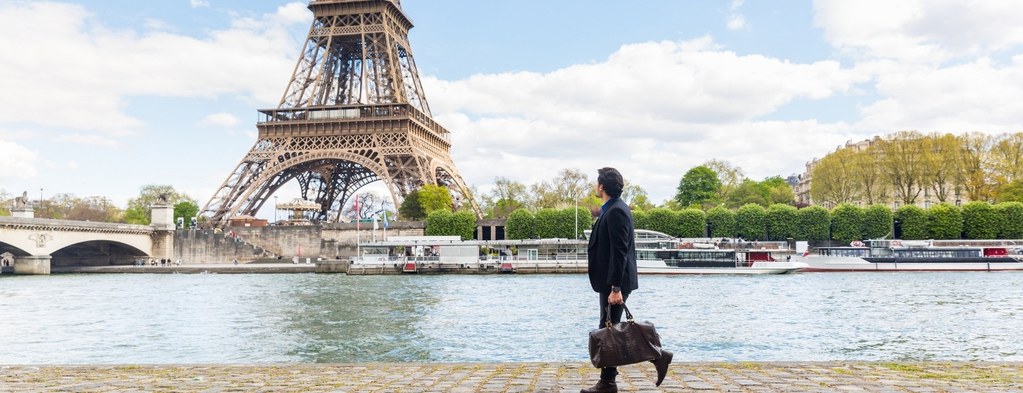 Person carrying a travel bag while walking along a riverside walkway, with the Eiffel Tower and riverboats in the background.