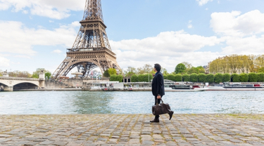 Une personne portant un sac de voyage qui marche le long d’une promenade au bord de la rivière, avec la Tour Eiffel et des bateaux de croisière en arrière-plan.