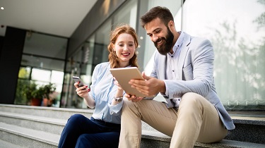 Image of two people sitting on steps outside a building, one is holding a mobile phone, the other is holding a tablet.