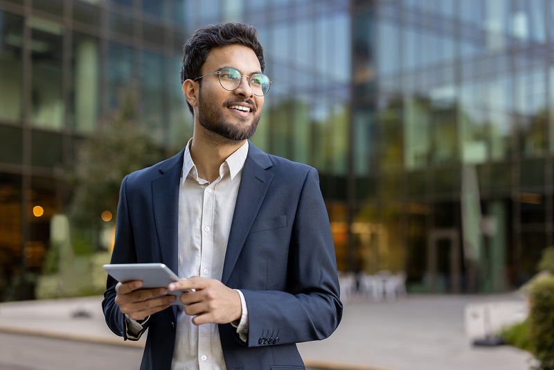 Person in a business suit holding a tablet outdoors in front of a modern glass office building.
