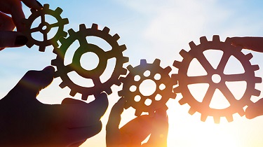 Four hands, each holding a gear, work together to move forward against a sunlit sky.