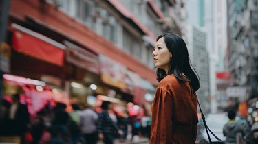 A woman stands on a busy street, surrounded by tall buildings and looking pensive.