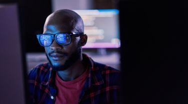 Un homme assis dans une pièce sombre devant son ordinateur, les lignes de code se reflétant sur ses lunettes.