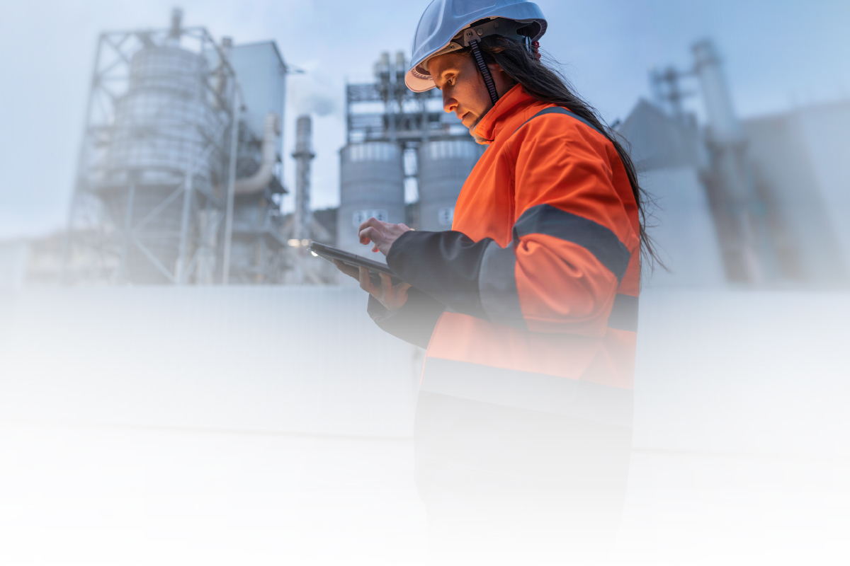 Woman wearing a hard hat using a tablet at an industrial facility, reviewing operations and supply chain information in a manufacturing environment.