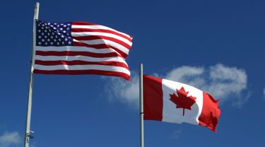 Les drapeaux des États-Unis et du Canada flottant au vent devant un ciel bleu et des nuages.