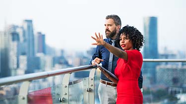 Businesswoman and businessman on a building rooftop describing something they see in the distance.