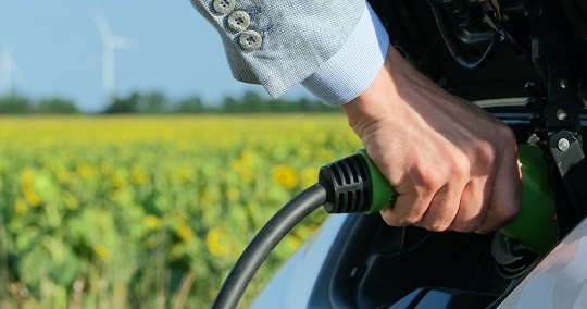 Man charging electric car with farm field and wind turbine in the background.