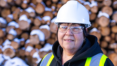 Female industrial worker with white hard hat and yellow safety vest standing in front of a blurry wood pile.