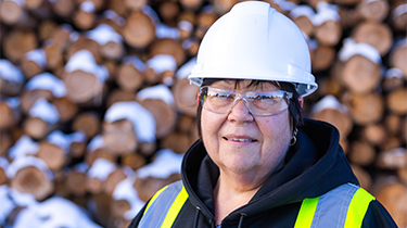 Ouvrière portant un casque de protection blanc et un gilet de sécurité jaune devant une pile de bois aux contours flous.