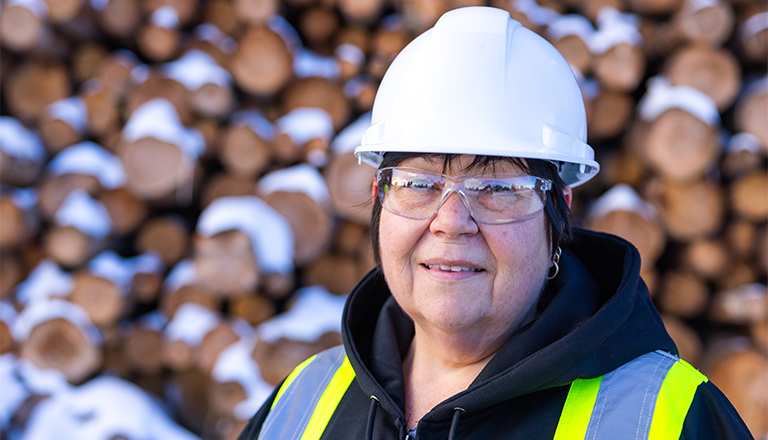 Ouvrière portant un casque de protection blanc et un gilet de sécurité jaune devant une pile de bois aux contours flous.