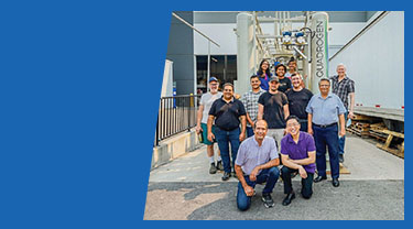 A group of Quadrogen employees stands in front of a biogas clean-up system near a warehouse. 