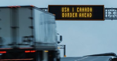 Transport truck passes under USA-Canada Border ahead sign on highway.
