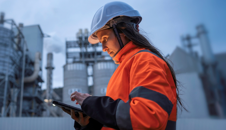 Personne portant un casque de sécurité et utilisant une tablette dans une installation industrielle pour consulter des informations sur les activités et la chaîne d’approvisionnement.