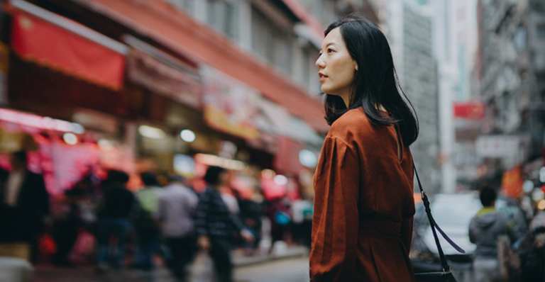 A woman stands on a busy street, surrounded by tall buildings and looking pensive.