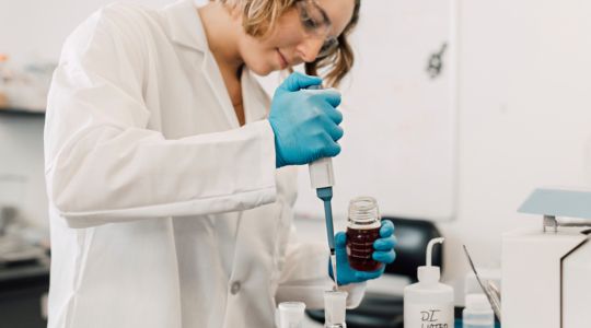 A woman wearing a lab coat and safety glasses transferring liquid from a beaker into an Erlenmeyer flask in a laboratory.