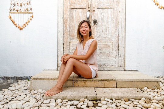 Joy Yap sits on a step of a home surrounded in natural wood, stone