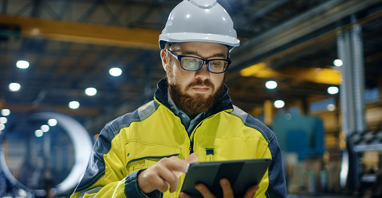 An engineer wearing a yellow hardhat in his factory is typing on his tablet.