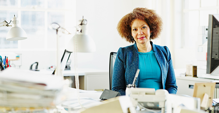 A woman sitting at her in front of a pill of paper is taking a pause in her busy day.