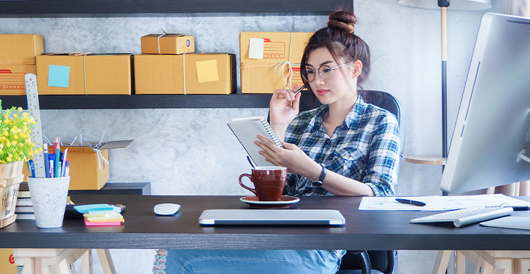 A woman at work is consulting her notes while drinking her coffee. 