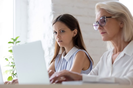 Young woman peeks at her employer’s computer screen with suspicion. 