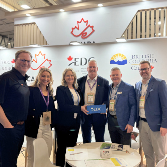 Group of people standing together and smiling at a trade show booth with visible logos for EDC, Canada, and British Columbia.