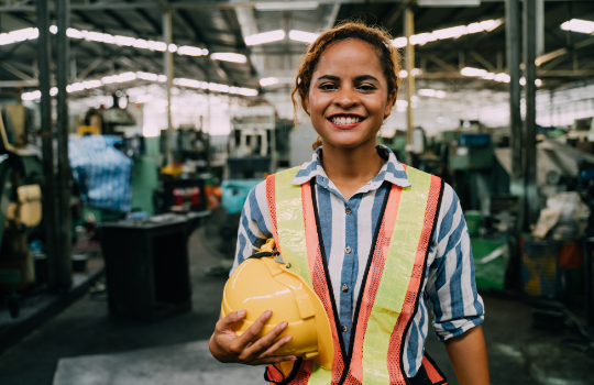 Portrait of Female Engineer wearing safety protective vest while standing front of production line at industrial manufacturing facility.
